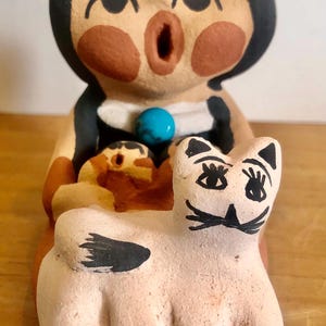 May include: A ceramic figurine of a seated person holding a cat. The figure has black hair, brown skin, and an open-mouthed expression. The cat is light pink with black markings. A turquoise bead is on the figure's chest.