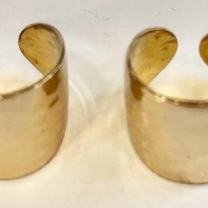 Puede incluir: Dos anillos de puño dorados de extremo abierto. Los anillos tienen una superficie lisa y pulida y una forma redondeada. La abertura permite un fácil uso. Estos anillos son un accesorio simple pero elegante.