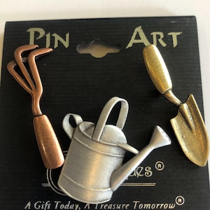 May include: A set of three garden-themed pins. The pins include a copper-colored rake, a silver watering can, and a gold-colored trowel. The pins are on a black card with the words "Pin Art" and "A Gift Today, A Treasure Tomorrow."