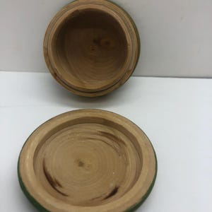 Op de afbeelding: Een kleine, open houten doos met een deksel. De doos en het deksel zijn gemaakt van lichtgekleurd hout met zichtbare nerfpatronen. De rand van de doos en het deksel zijn groen geverfd. De doos is rond en heeft een eenvoudig, natuurlijk ontwerp.