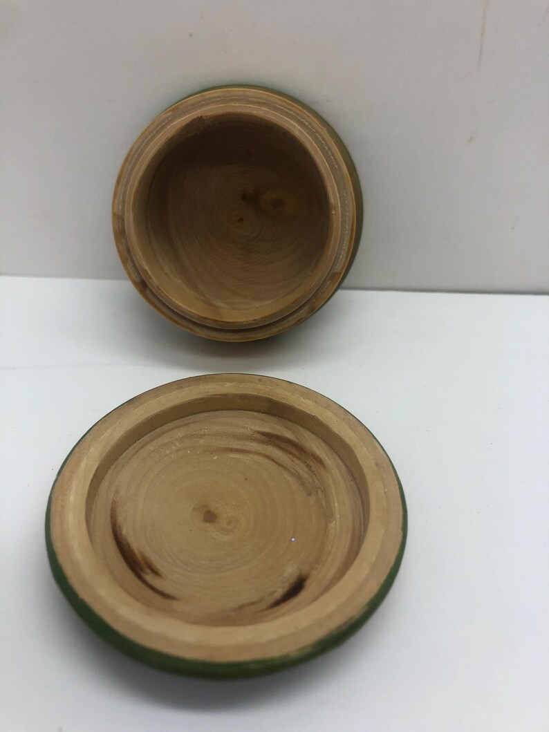 Op de afbeelding: Een kleine, open houten doos met een deksel. De doos en het deksel zijn gemaakt van lichtgekleurd hout met zichtbare nerfpatronen. De rand van de doos en het deksel zijn groen geverfd. De doos is rond en heeft een eenvoudig, natuurlijk ontwerp.