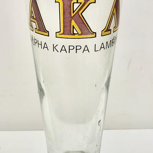 Op de afbeelding: Helder glazen pilsnerglas met een brede voet en een slanke, taps toelopende body. Het glas heeft de letters "AKA" in kastanjebruin met gele contouren en de woorden "Alpha Kappa Lambda" eronder.