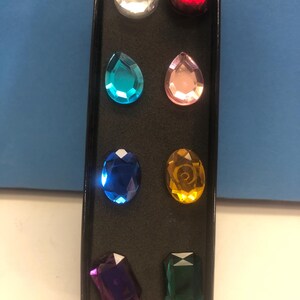May include: A black rectangular display box containing eight faceted acrylic gemstones in various shapes and colors, including clear, red, teal, light pink, blue, yellow, purple, and green, arranged in two vertical columns on a black foam insert.