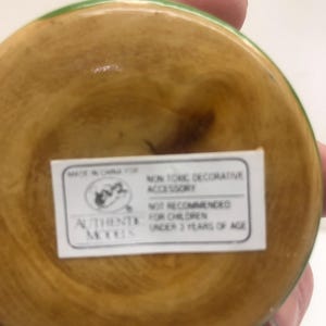 Op de afbeelding: Een houten decoratief accessoire met een lichtbruin interieur en een groene rand. De onderkant van het accessoire heeft een wit label met de tekst "NON TONIC DECORATIVE ACCESSORY" en "NOT RECOMMENDED FOR CHILDREN UNDER 3 YEARS OF AGE."
