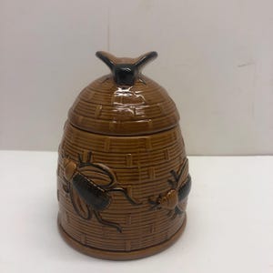 May include: Brown ceramic beehive-shaped jar with a lid and two black insect designs. The jar has a textured, woven pattern and a small handle on top. A decorative kitchen item.