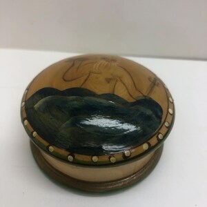 Op de afbeelding: Een ronde houten doos met een deksel met een geschilderde sc&egrave;ne van een figuur en donkerblauwe golven. De doos heeft een lichtbruine basis en een decoratieve rand met kleine witte stippen. Het ontwerp van het deksel toont een figuur en donkerblauwe golven.
