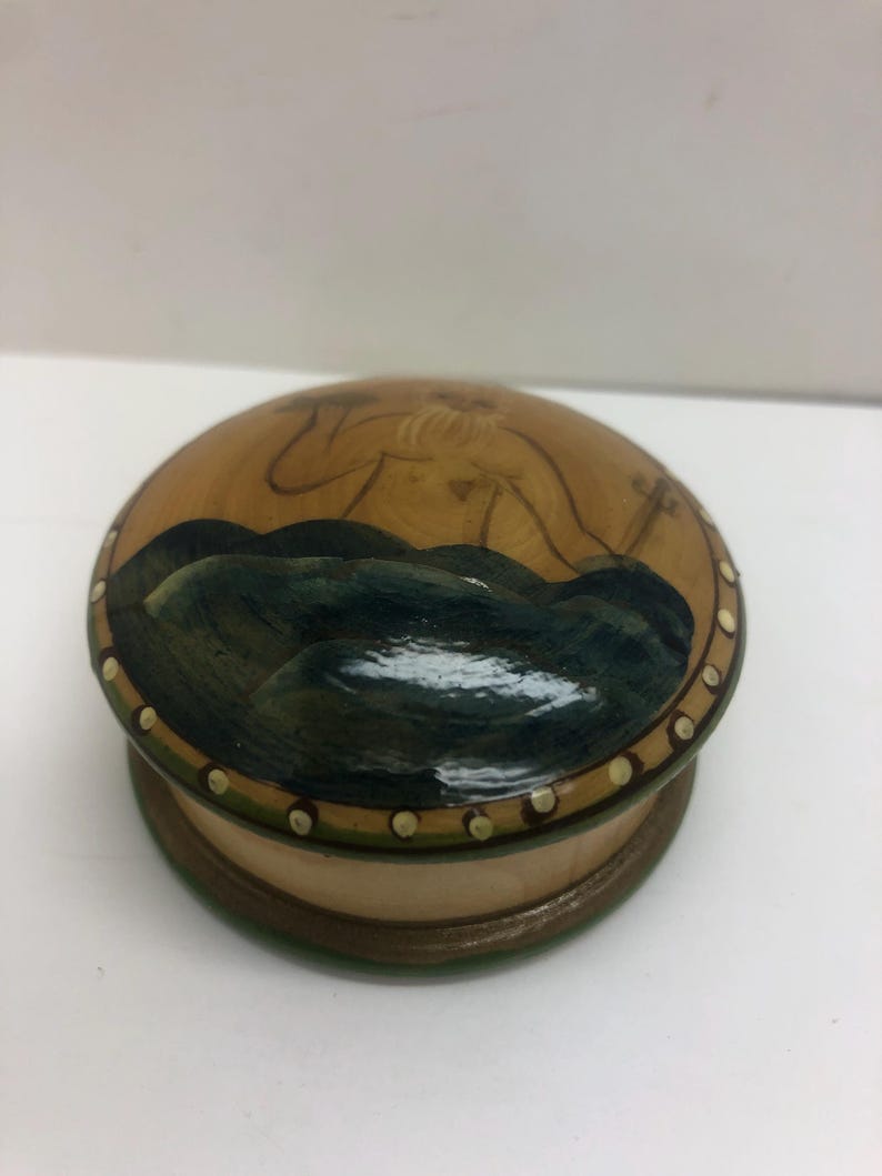 Op de afbeelding: Een ronde houten doos met een deksel met een geschilderde sc&egrave;ne van een figuur en donkerblauwe golven. De doos heeft een lichtbruine basis en een decoratieve rand met kleine witte stippen. Het ontwerp van het deksel toont een figuur en donkerblauwe golven.
