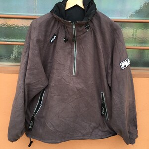 Op de afbeelding: Een bruine Fila anorakjas met een zwarte capuchon en een ritssluiting. De jas heeft het Fila-logo op de borst en mouw. Het heeft twee zakken met ritssluiting en een trekkoord aan de onderkant. De jas hangt aan een houten hanger.