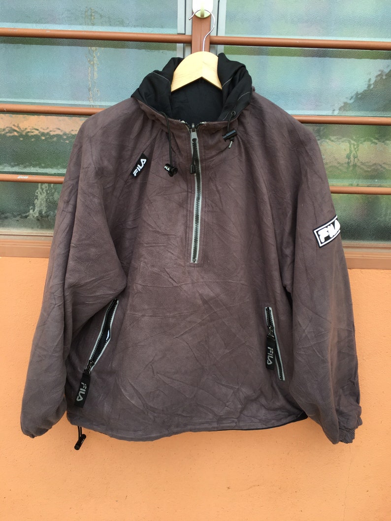 Op de afbeelding: Een bruine Fila anorakjas met een zwarte capuchon en een ritssluiting. De jas heeft het Fila-logo op de borst en mouw. Het heeft twee zakken met ritssluiting en een trekkoord aan de onderkant. De jas hangt aan een houten hanger.