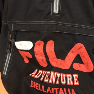 Op de afbeelding: Zwarte anorakjas met een groot rood en wit Fila-logo. De jas heeft een borstzak met ritssluiting, een halve ritssluiting en de woorden "ADVENTURE BIELLA ITALIA" in witte letters.