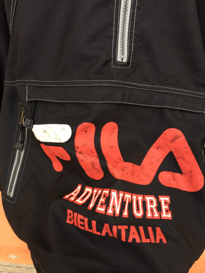 Op de afbeelding: Zwarte anorakjas met een groot rood en wit Fila-logo. De jas heeft een borstzak met ritssluiting, een halve ritssluiting en de woorden "ADVENTURE BIELLA ITALIA" in witte letters.