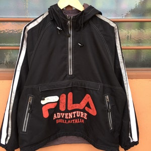 Op de afbeelding: Zwarte anorakjas met capuchon, witte strepen op de mouwen en het FILA-logo in rood en wit. De jas heeft een halve ritssluiting, twee zakken met ritssluiting en de tekst "ADVENTURE BIELLITALIA".