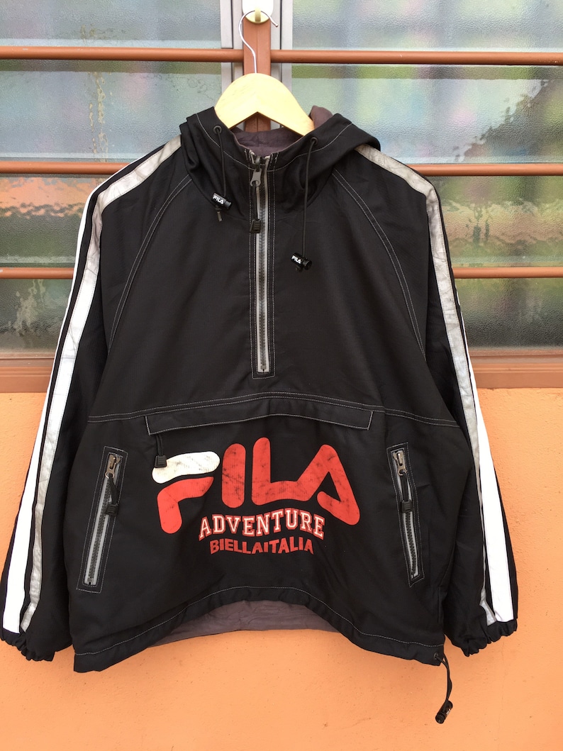 Op de afbeelding: Zwarte anorakjas met capuchon, witte strepen op de mouwen en het FILA-logo in rood en wit. De jas heeft een halve ritssluiting, twee zakken met ritssluiting en de tekst "ADVENTURE BIELLITALIA".