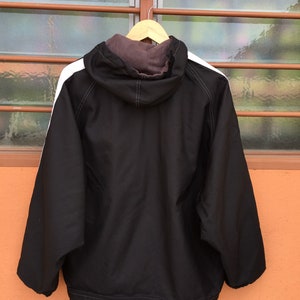 Op de afbeelding: Zwarte jas met capuchon en witte strepen op de schouders, hangend aan een houten hanger. De jas heeft een trekkoord aan de onderkant en een bruine voering in de capuchon. De achtergrond is een raam met een bruin frame.