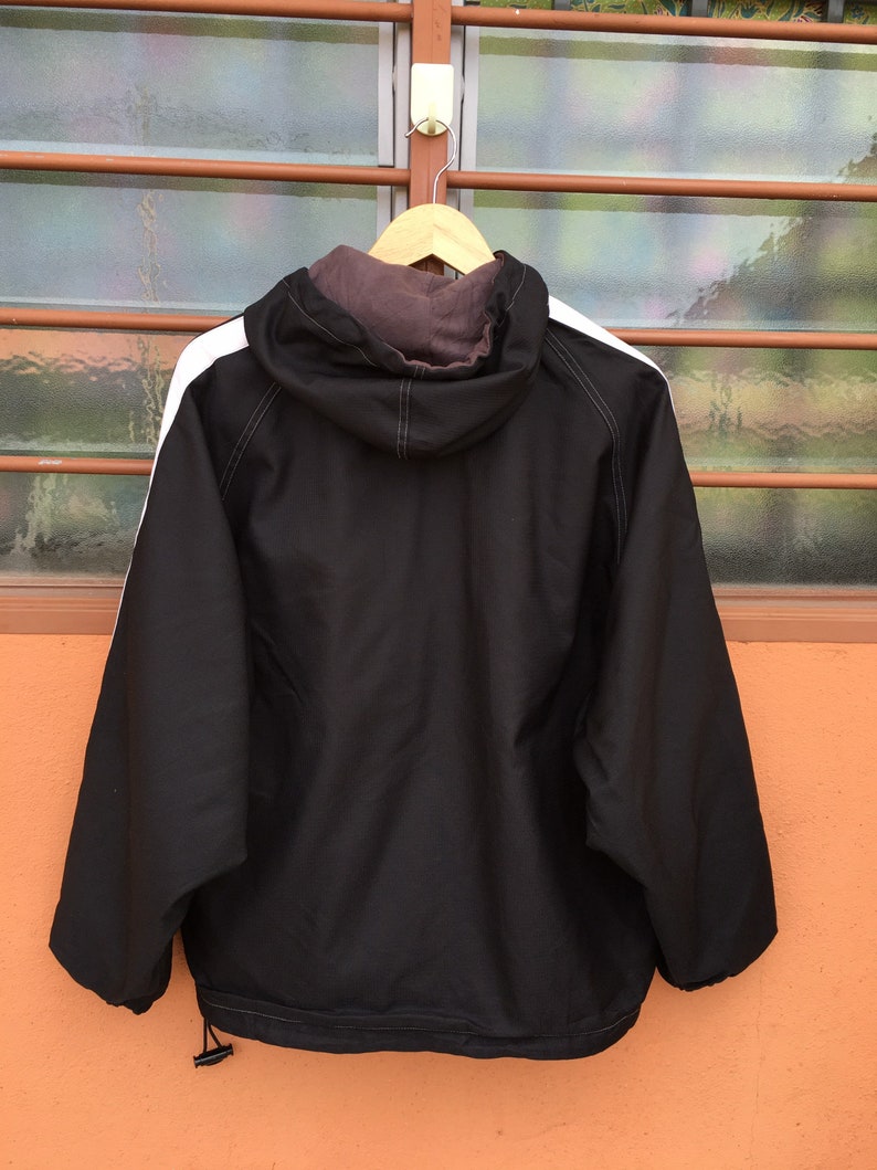 Op de afbeelding: Zwarte jas met capuchon en witte strepen op de schouders, hangend aan een houten hanger. De jas heeft een trekkoord aan de onderkant en een bruine voering in de capuchon. De achtergrond is een raam met een bruin frame.