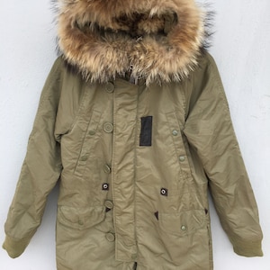 Może przedstawiać: Zielona parka z kapturem obszytym brązowym futrem. Kurtka zapinana na zamek błyskawiczny i wiele guzików. Kurtka ma dwie kieszenie z przodu i naszywkę na lewym rękawie.