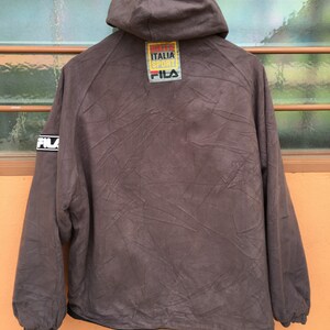Op de afbeelding: Bruine jas met capuchon met het "FILA"-logo op de mouw en rug. De jas heeft een trekkoord aan de onderkant en een label met de tekst "BUTTER ITALIA SPORT FILA". De jas is gemaakt van een textuurrijk materiaal.