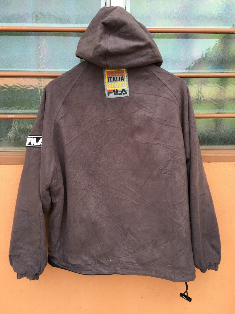 Op de afbeelding: Bruine jas met capuchon met het "FILA"-logo op de mouw en rug. De jas heeft een trekkoord aan de onderkant en een label met de tekst "BUTTER ITALIA SPORT FILA". De jas is gemaakt van een textuurrijk materiaal.