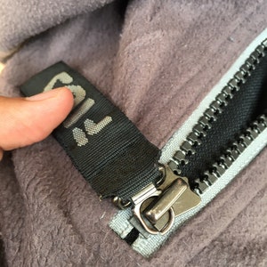 Op de afbeelding: Close-up van een grijze fleecejas met een zwarte rits en een zwart label met het woord "FILA" in het wit. De rits is zilverkleurig en de stof is zacht en getextureerd.