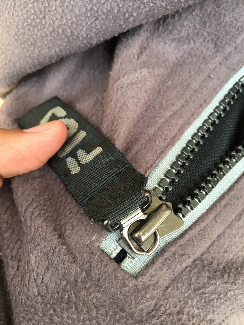 Op de afbeelding: Close-up van een grijze fleecejas met een zwarte rits en een zwart label met het woord "FILA" in het wit. De rits is zilverkleurig en de stof is zacht en getextureerd.