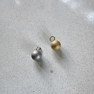 Op de afbeelding: Twee bolvormige bedels, één in een metallic zilveren afwerking en één in een metallic gouden afwerking. Elke bedel heeft een getextureerd, glinsterend oppervlak en is voorzien van een klein rond ringetje aan de bovenkant.