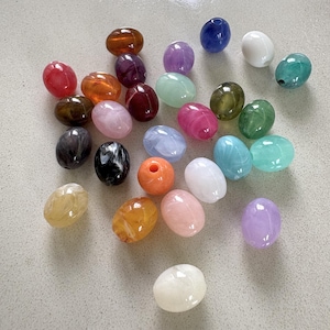 Könnte beinhalten: Eine Sammlung ovaler Perlen in verschiedenen Farben, darunter Rot, Orange, Gelb, Grün, Blau, Violett, Rosa, Weiß, Schwarz und Braun. Jede Perle hat eine glatte, polierte Oberfläche mit einem marmorierten oder gewirbelten Erscheinungsbild.