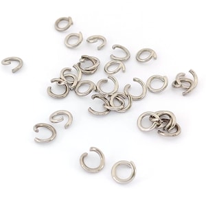 Op de afbeelding: Een verzameling kleine, zilverkleurige sprongringen verspreid over een wit oppervlak. De ringen zijn open, met een C-vormig ontwerp, en worden waarschijnlijk gebruikt voor het maken van sieraden of knutselen. Het metaal heeft een glanzende afwerking.