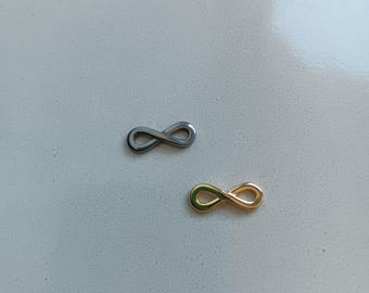 May include: Two infinity symbol charms. One is silver-colored, and the other is gold-colored. The charms are small and likely used for jewelry making or crafting. They are placed on a light gray surface.