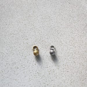 Op de afbeelding: Twee kleine bolvormige bedels, één goudkleurig en één zilverkleurig, elk met een getextureerd, glinsterend oppervlak en een kleine ronde ring aan de bovenkant om op te hangen of aan sieraden te bevestigen.