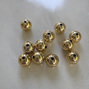 May include: A collection of ten spherical beads with a polished gold-tone finish. Each bead features a smooth, reflective surface and a single centered hole for threading, arranged in a loose cluster on a light-colored surface.