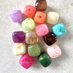 Könnte beinhalten: Eine Sammlung quadratischer Perlen mit abgerundeten Ecken, die ein marmoriertes, halbtransparentes Finish in verschiedenen Farben wie Lila, Pink, Orange, Braun, Creme, Grün und Blau aufweisen, jede mit einem zentralen Loch zum Auffädeln.