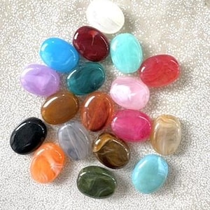 Könnte beinhalten: Eine Sammlung ovaler Cabochons in verschiedenen Farben, darunter Weiß, Rot, Blau, Grün, Violett, Braun, Schwarz, Orange, Pink und Beige. Jedes Stück hat eine glatte, polierte Oberfläche mit einer marmorierten oder gewirbelten Textur.