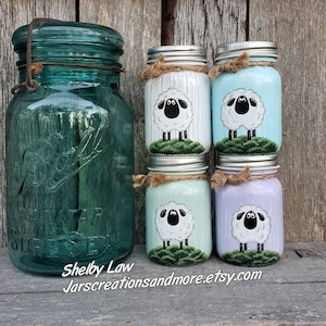 Peut inclure: Quatre bocaux en verre avec des moutons peints. Les bocaux sont peints dans des tons de bleu, vert et violet. Les bocaux sont décorés de ficelle et ont des couvercles argentés. Les bocaux sont posés sur une surface en bois. Le texte "Shelby Law Jarscreationsandmore.etsy.com" est visible en bas de l'image.