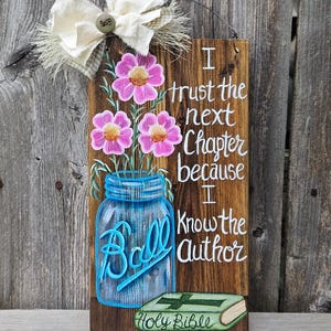 May include: Wooden sign with a rustic aesthetic. It features a painted blue mason jar with pink flowers, and the text "I trust the next chapter because I know the Author." A small green Bible is also depicted.