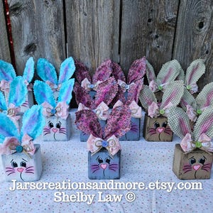 May include: A collection of 10 handmade wooden bunny blocks decorated with fabric ears and bows. The bunnies are painted in various colors, including blue, pink, and brown. The blocks are a fun and festive way to decorate for Easter.