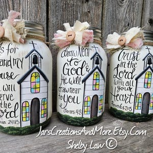 May include: Three hand-painted mason jars with church designs and inspirational quotes. Each jar features a white church with colourful stained-glass windows and a cross. The jars are decorated with fabric and buttons.