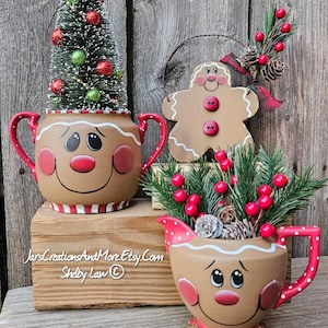 May include: Two tan ceramic containers with painted gingerbread faces, red handles, and white icing details. One holds a small flocked Christmas tree with red and green ornaments, the other holds pine branches and red berries. A gingerbread ornament is included.