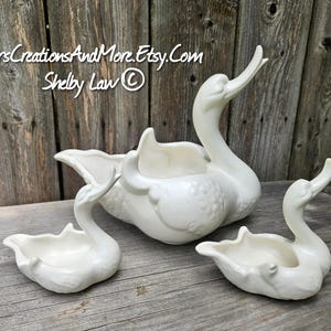 May include: A set of three white ceramic swan-shaped dishes. The largest swan features an upright neck and a hollow body, while the two smaller swan dishes have lower profiles. The text JarsCreationsAndMore.Etsy.Com Shelby Law is visible.