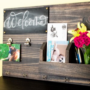 May include: A dark brown wooden wall organizer with a chalkboard that says "Welcome", two photo clips, a small shelf with a vase of yellow and pink flowers, and three hooks.