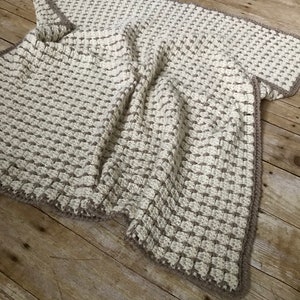 May include: A crocheted throw blanket in cream and brown with a textured stitch pattern. The blanket is folded and lying on a wooden surface.