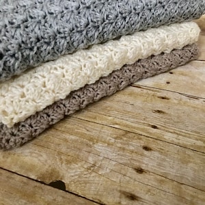 May include: Three crocheted blankets stacked on a wooden surface. The top blanket is gray, the middle blanket is white, and the bottom blanket is brown.