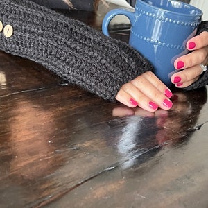 May include: A close-up of a person's hand wearing a gray knitted fingerless glove, holding a blue ceramic mug with a beaded rim. The mug is filled with a warm beverage.