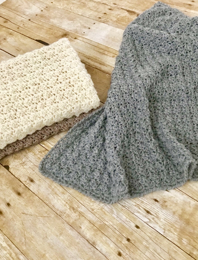 May include: Two crocheted blankets, one in a light beige color and the other in a dark gray color, are folded and placed on a wooden surface.