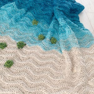 May include: A handmade crochet blanket depicting a beach scene. The blanket features a gradient of blue and turquoise representing the ocean, transitioning to a beige colour for the sand. Small green turtle appliqués are scattered across the sand.