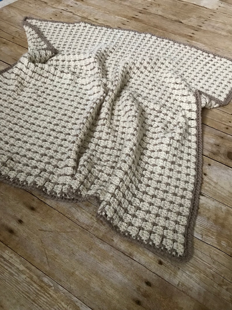 May include: A crocheted throw blanket in cream and brown with a textured stitch pattern. The blanket is folded and lying on a wooden surface.