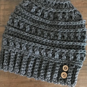 May include: A grey crocheted beanie with two brown buttons. The beanie has a textured pattern and is made with a thick yarn.
