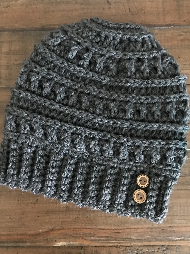 May include: A grey crocheted beanie with two brown buttons. The beanie has a textured pattern and is made with a thick yarn.