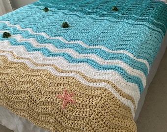 May include: A handmade crochet blanket with a beach theme. The blanket features a tan, white, and blue wave pattern, with a pink starfish and green accents. The blanket is displayed on a bed with white pillows.