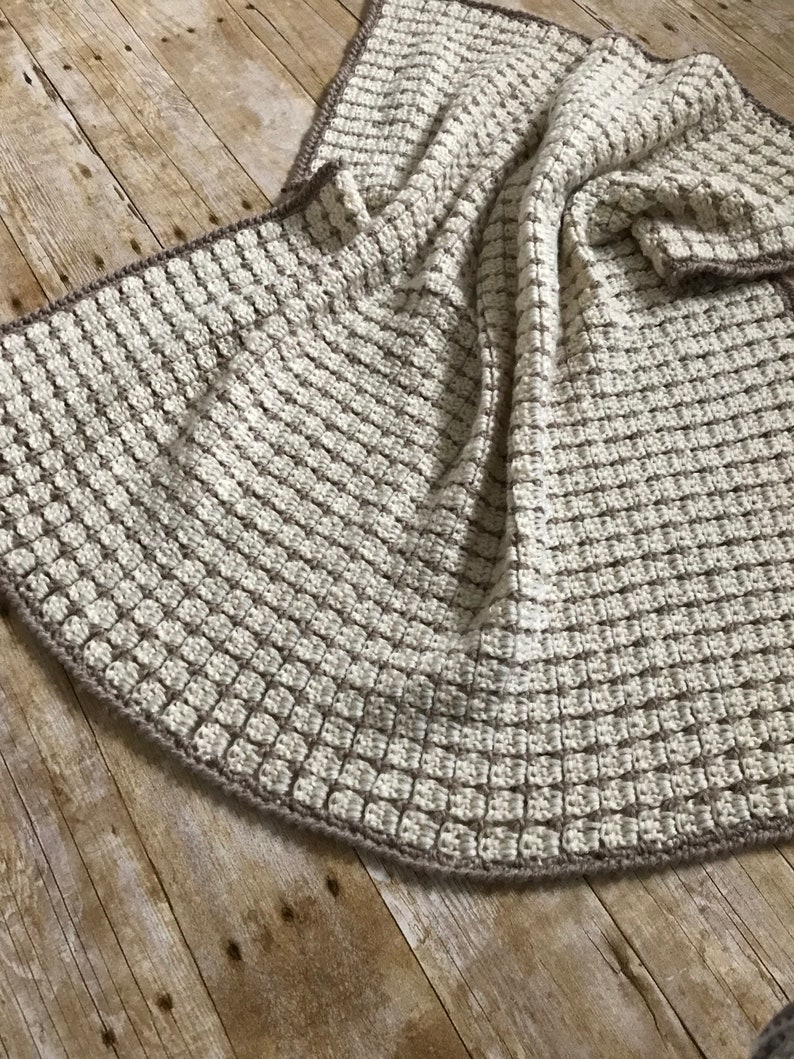 May include: A crocheted throw blanket in a cream and brown color scheme. The blanket is made with a textured stitch and has a crocheted border.
