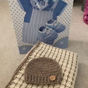 May include: A brown crocheted baby hat with a white button on top, resting on a white and brown crocheted blanket.